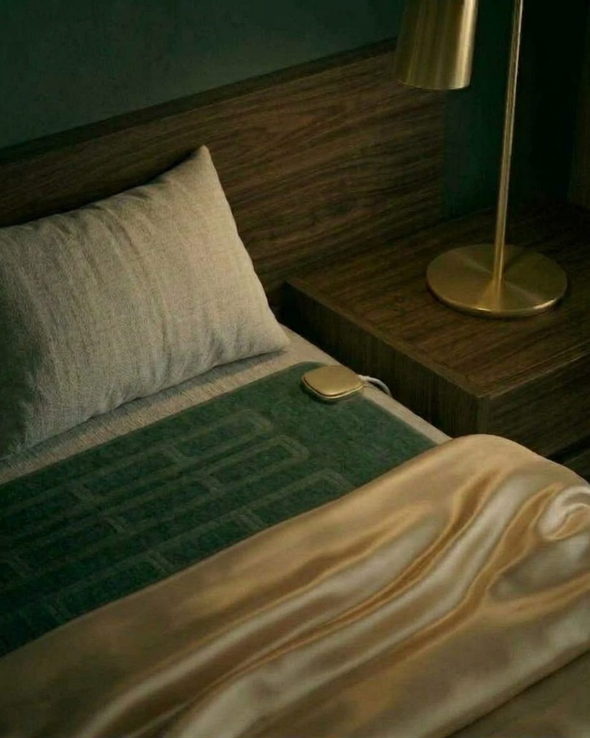Lonvi Pulse: a brushed champagne-gold palm device with a slim cable on dark green quilted bedding beside a brass lamp and walnut nightstand
